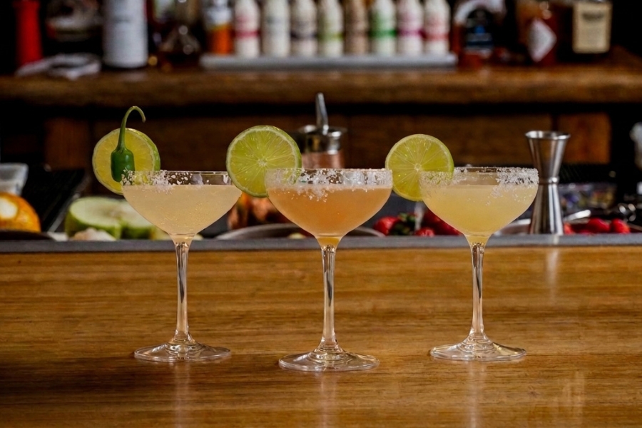 Three margarita cocktails with salted rims and lime wedges on a bar during happy hour.