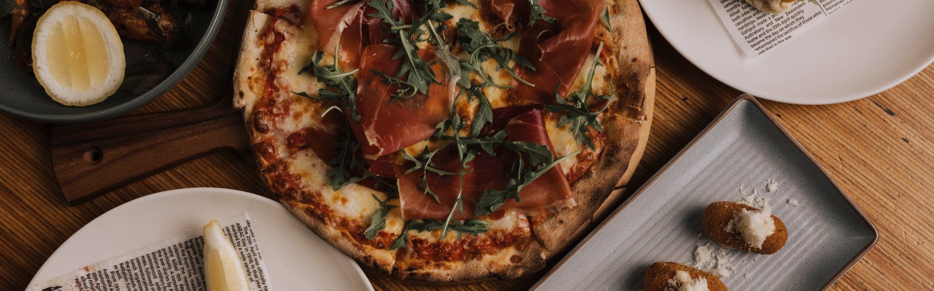 Wood-fired prosciutto pizza topped with rocket, served with sides and small plates on a dining table.