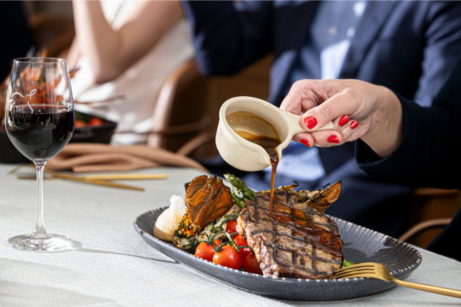 Diner pouring sauce over grilled steak at Ternary Restaurant, served with vegetables and red wine