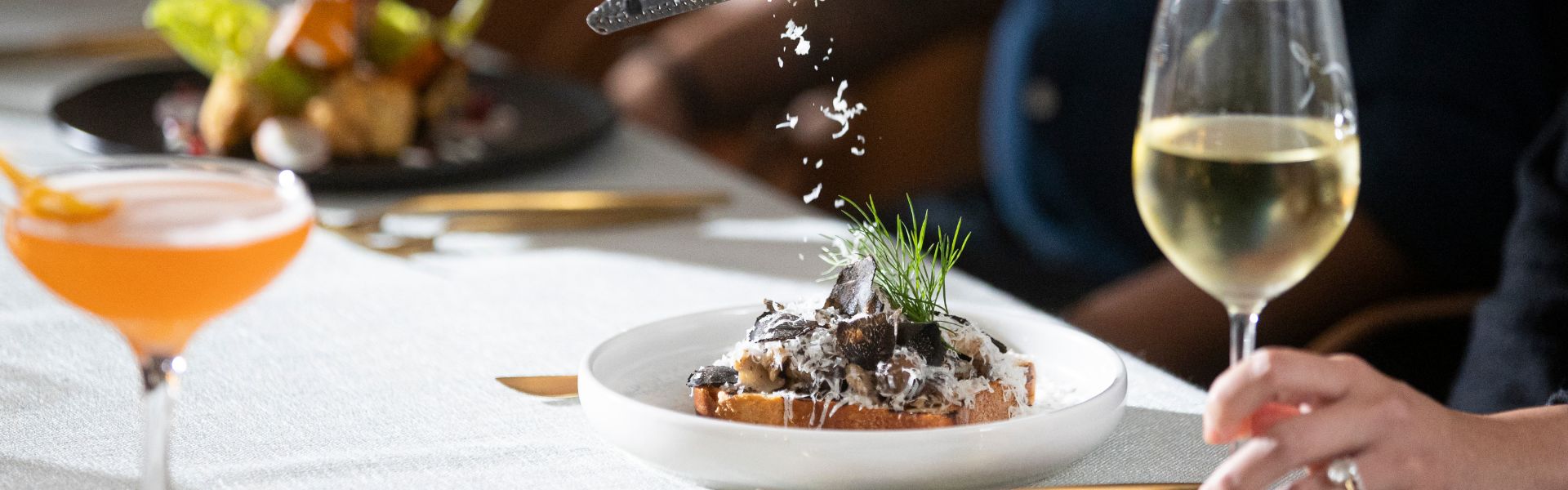 Truffle mushroom dish on toast being finished with shaved cheese, served with wine and a cocktail.